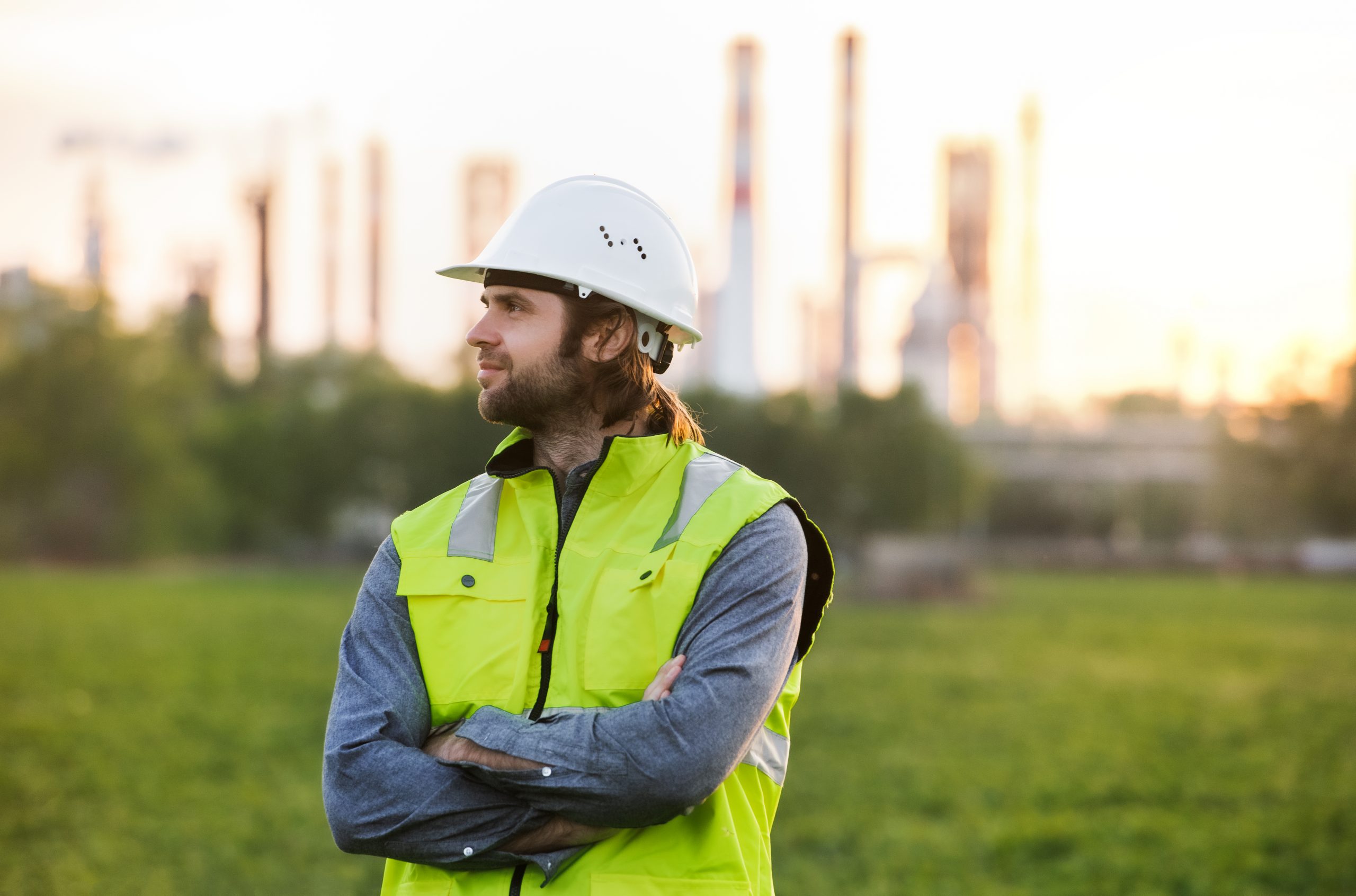 Young engineer standing outdoors by oil refinery, arms crossed.