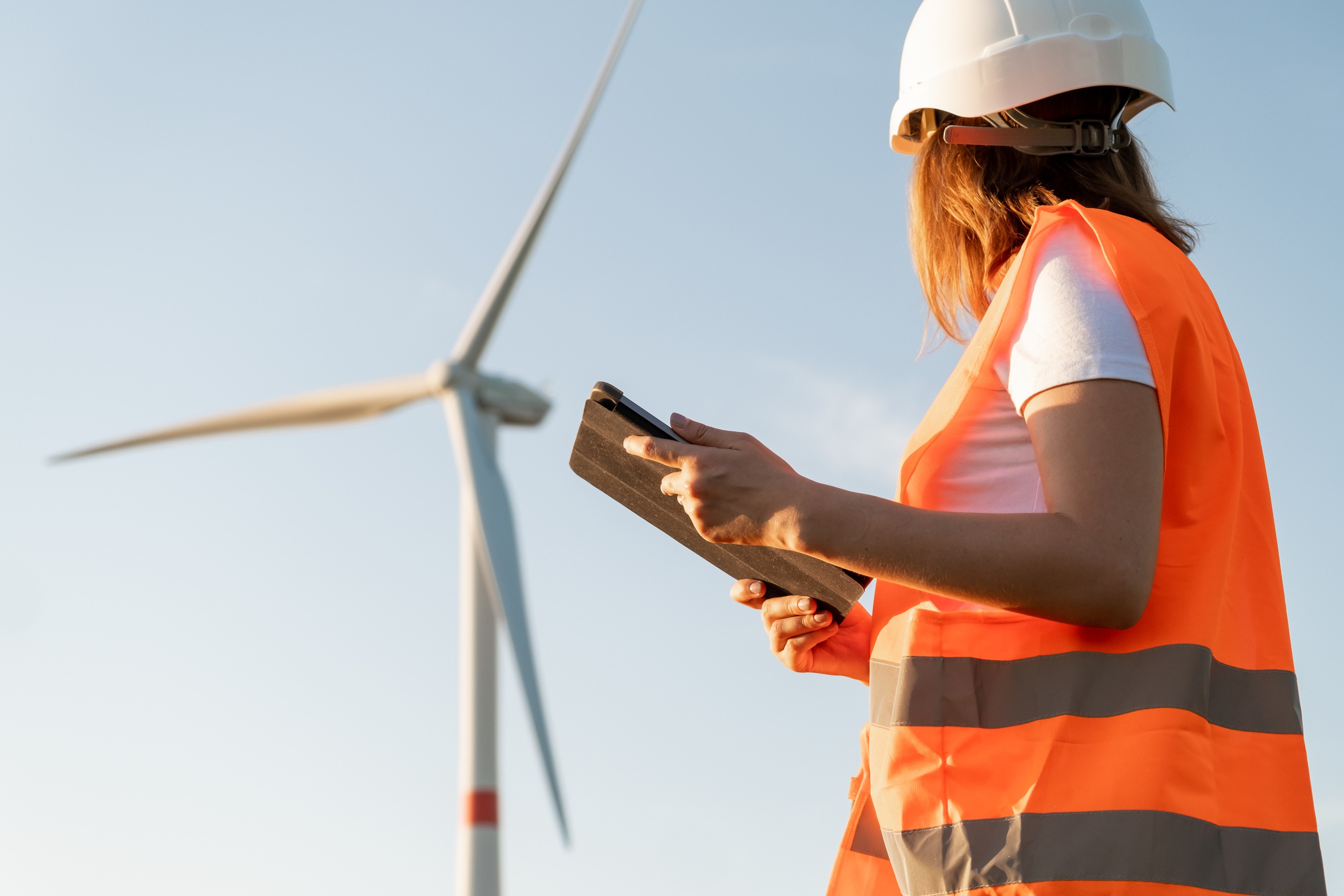 Female an engineer in a helmet and an orange vest controls the operation of a wind turbine renewable using a tablet on the sky background
