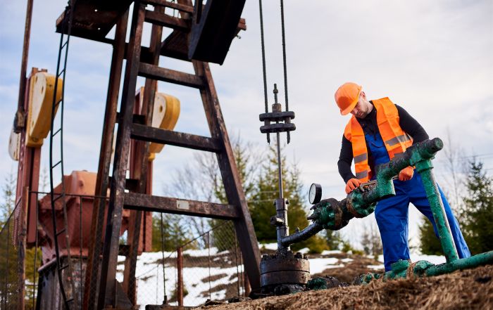 Oil worker in orange uniform and helmet working with a pipe wrench near an oil pump jack