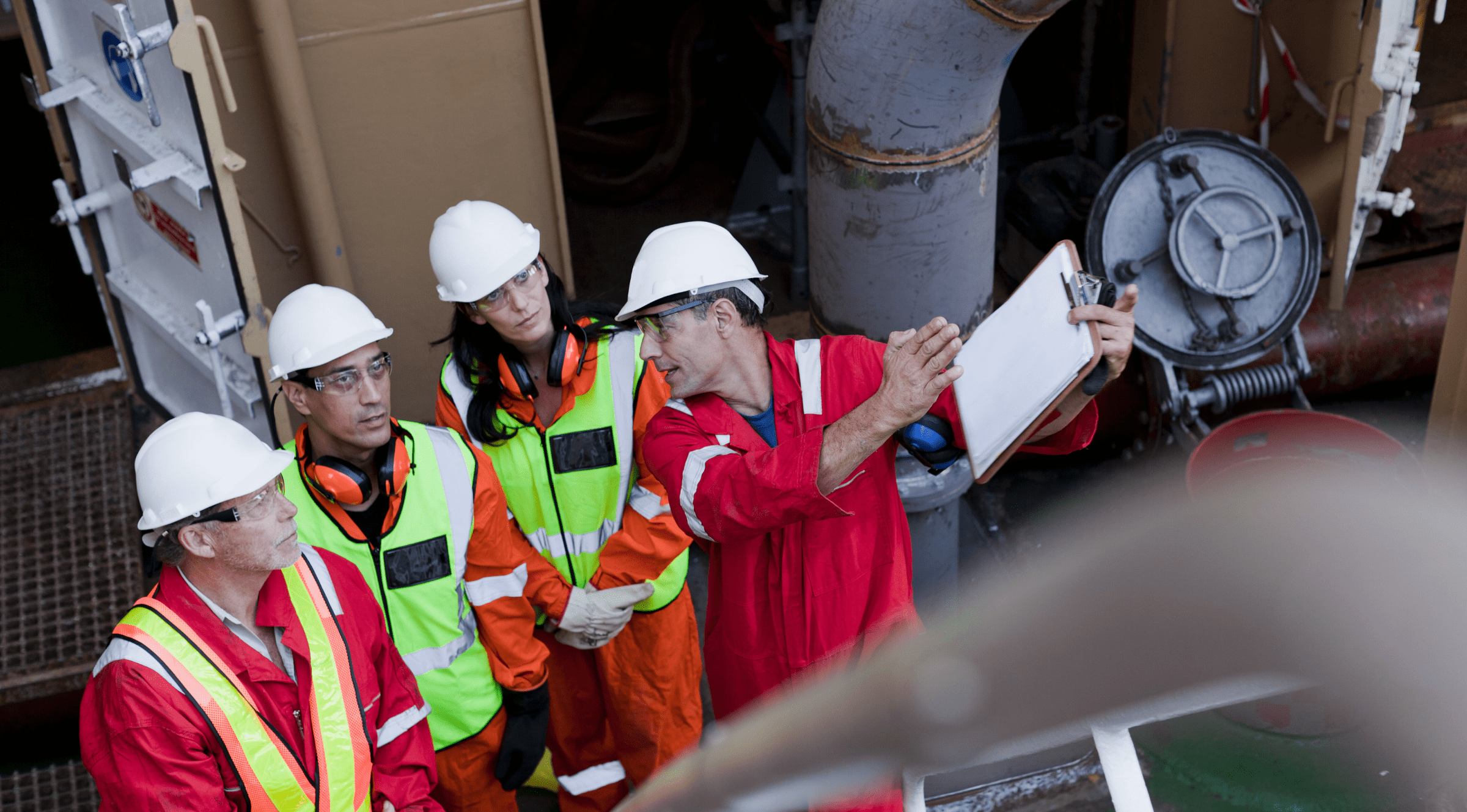 Four industrial inspectors wearing safety helmets and protective gear review equipment and discuss maintenance findings on an offshore facility.